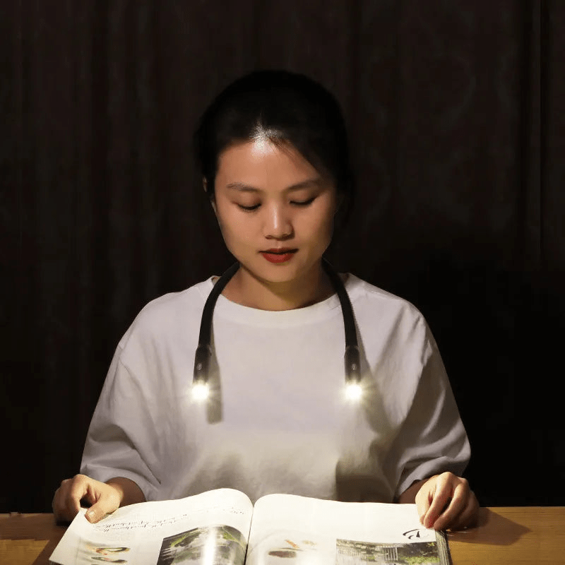 A person wearing a flexible, black Fitlybuy hands-free rechargeable neck reading light while reading a book in the dark.
