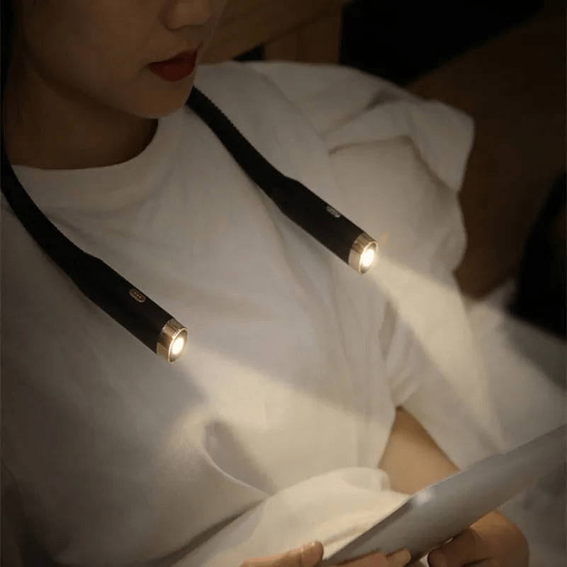 A person wears a black, flexible Fitlybuy FlexRead hands-free USB neck reading light while reading in bed.