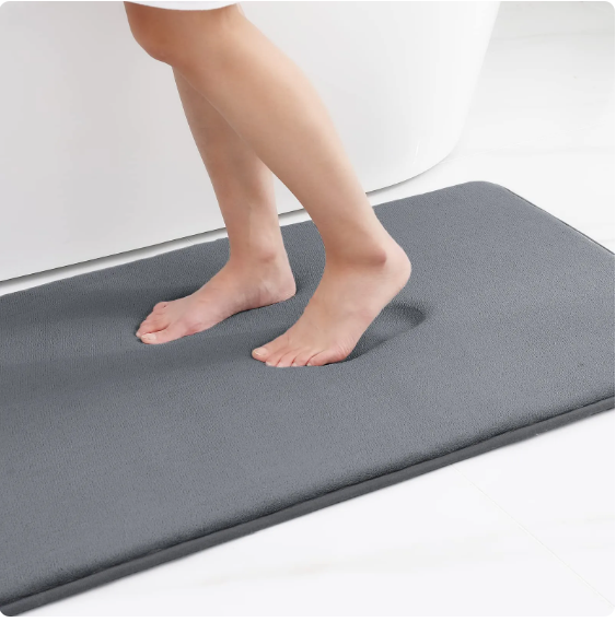 Bare feet step on a soft, grey Fitlybuy memory foam bath mat, showing an impression in the absorbent fabric.