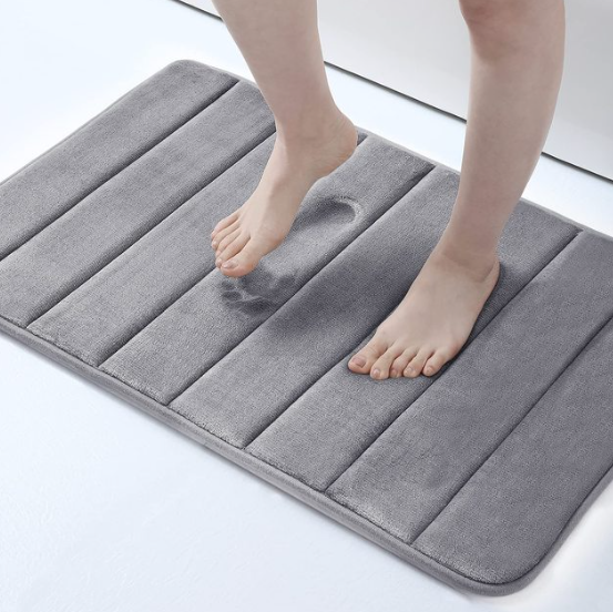 Person standing on a gray, striped Fitlybuy memory foam bath mat that shows a visible, absorbent footprint indentation.