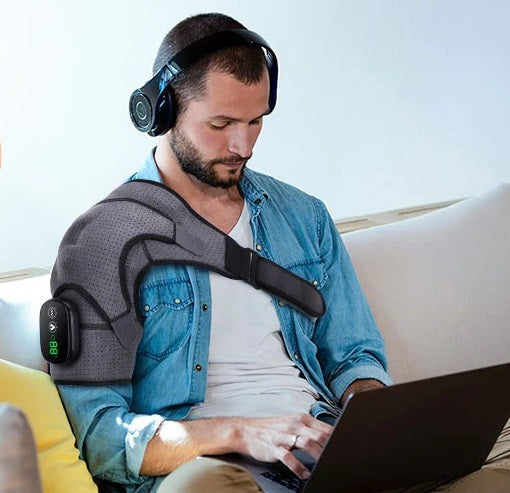 Man wearing a grey Fitlybuy heated shoulder compression brace with vibration therapy while working on a laptop.