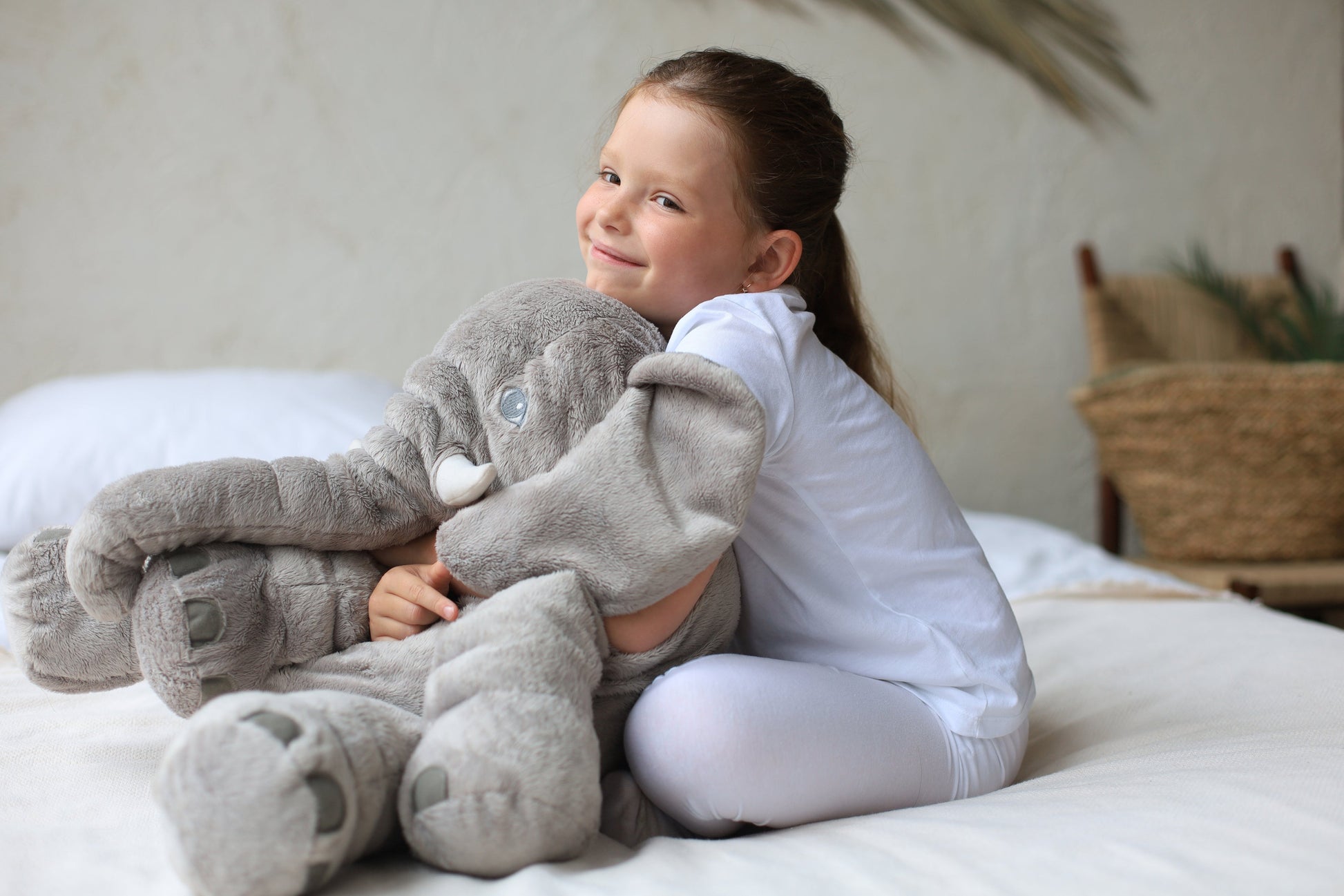 A young girl hugs a soft, grey Fitlybuy plush elephant pillow while sitting on a cozy, light-colored bed.