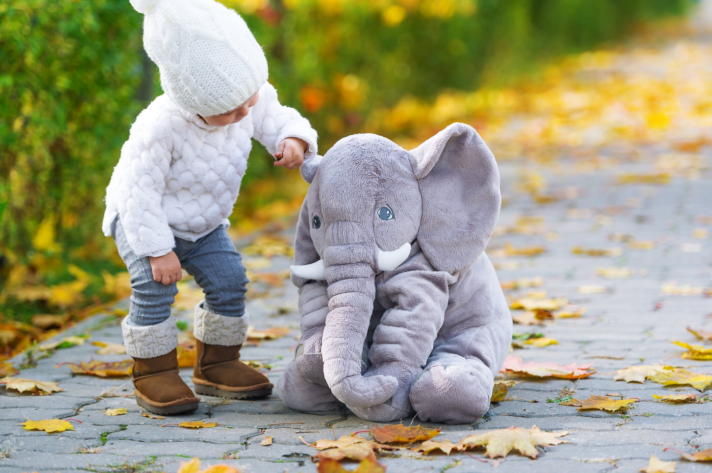 A toddler in a winter outfit touches a soft, gray Fitlybuy plush elephant pillow on an autumn path with fallen leaves.
