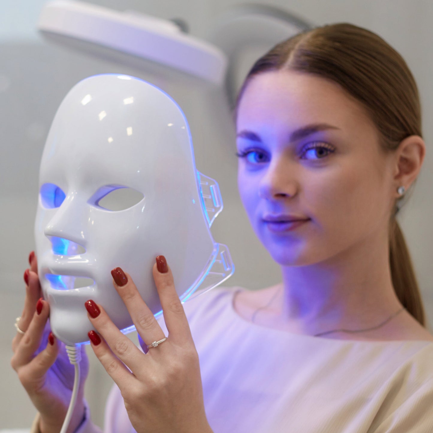 Woman holding a white Fitlybuy LED light therapy mask with blue light treatment in a clinical setting.