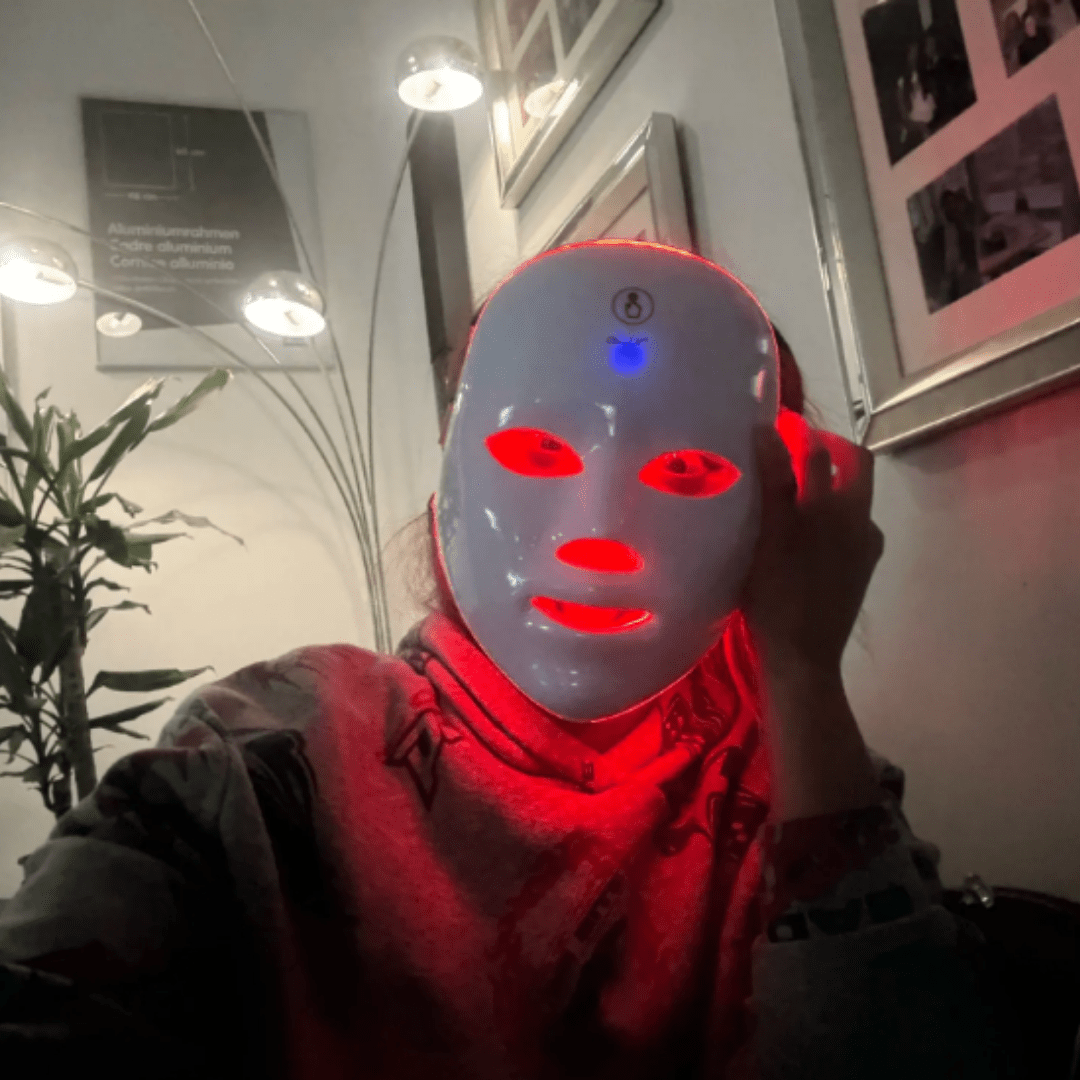Fitlybuy white LED light therapy mask with glowing red face light and neck strip, worn indoors by a user.