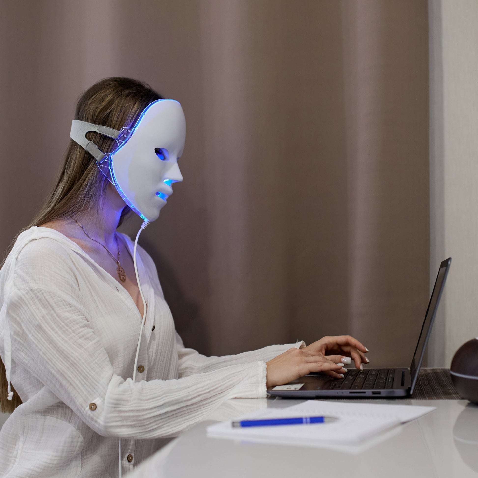 Woman wearing a white Fitlybuy red light LED mask with blue light, working on a laptop at a desk.