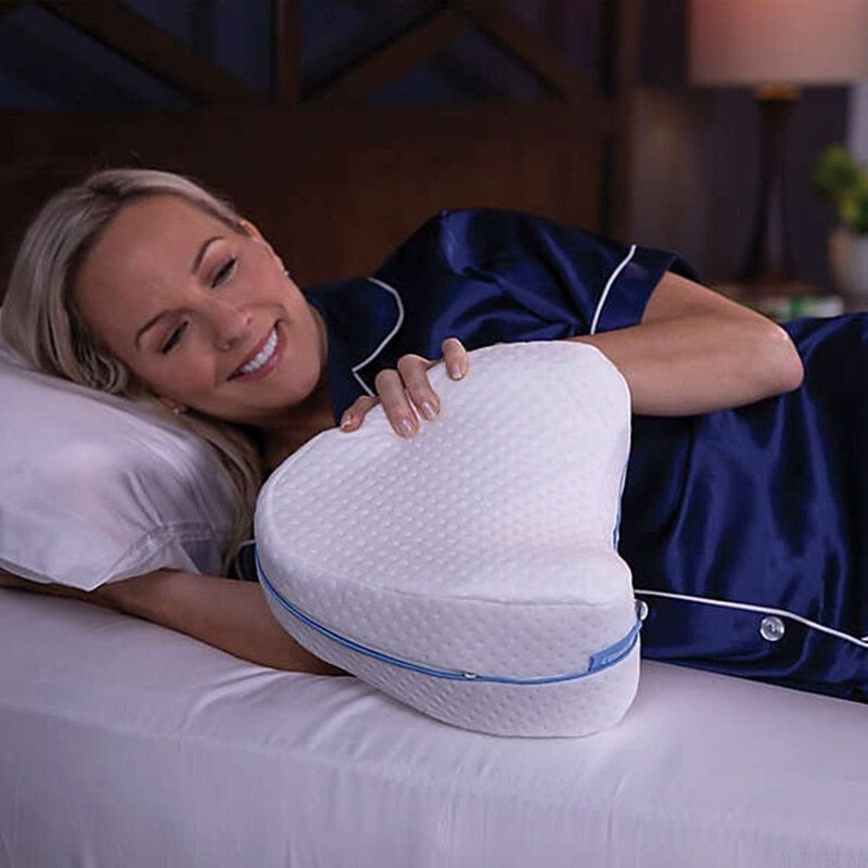 Woman in blue pajamas resting on a bed, holding a white, heart-shaped Fitlybuy memory foam knee pillow with blue trim.