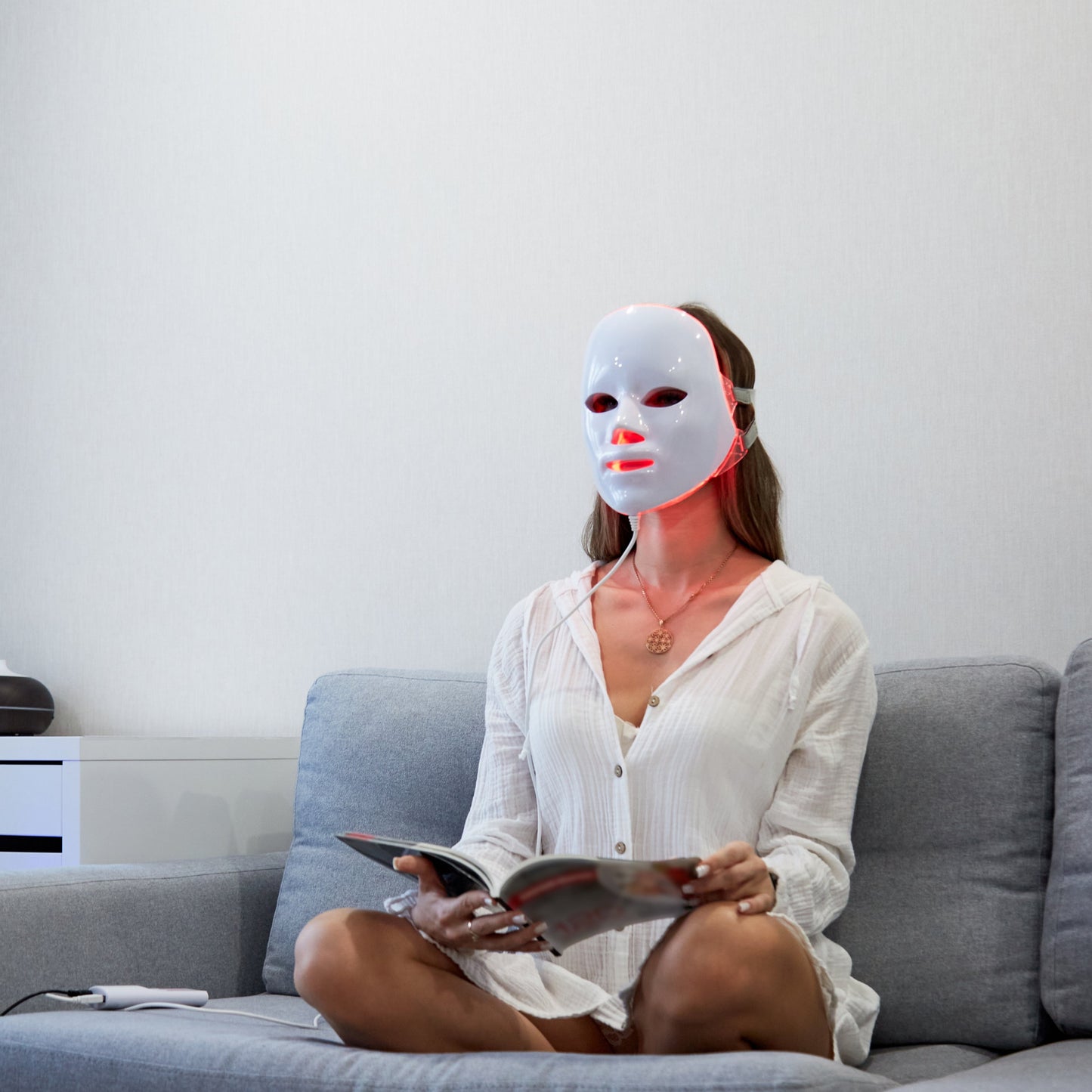 Woman on couch wearing white Fitlybuy red light LED mask for skincare while reading.