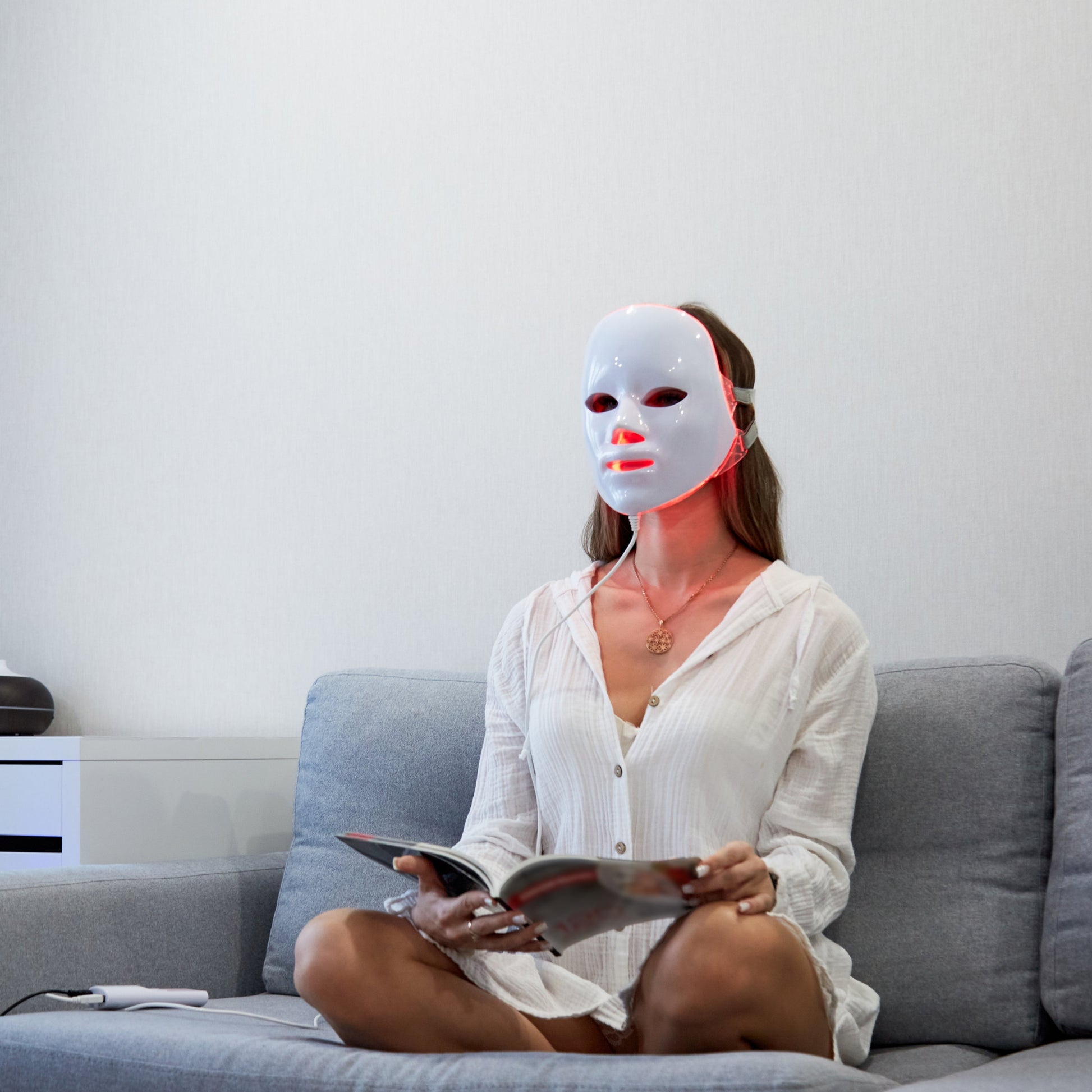 Woman on couch wearing white Fitlybuy red light LED mask for skincare while reading.