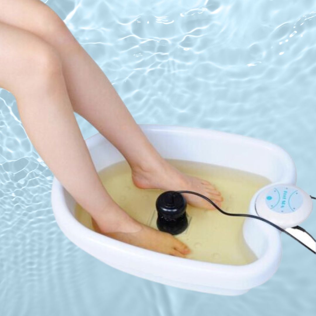White ionic foot spa basin with black detox array and control panel, feet submerged in yellow-tinted water.