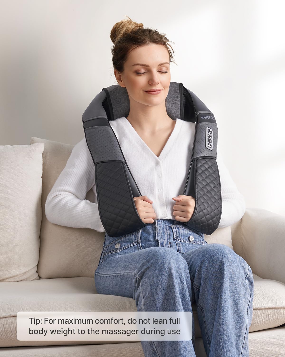 Woman sitting on a sofa wearing a grey U-shaped neck and shoulder massager with heat therapy and control buttons.
