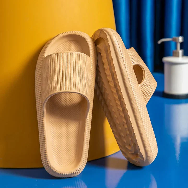 Tan Fitlybuy Cloud Slides with ComfortFoam textured soles, propped against a yellow background on a blue surface.