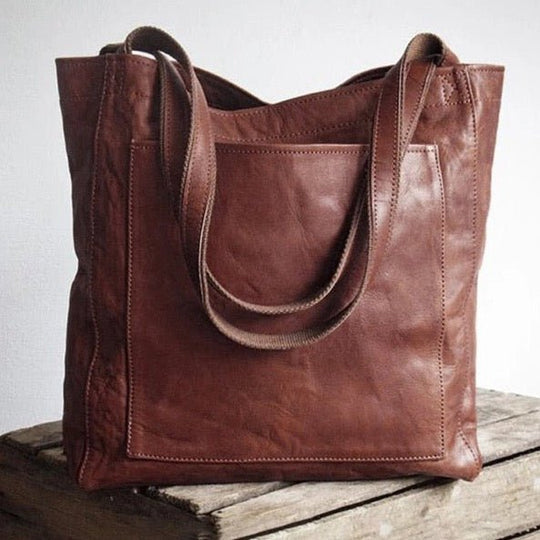 Brown leather Fitlybuy Eva tote bag with dual shoulder straps and a large front pocket, resting on a wooden crate.