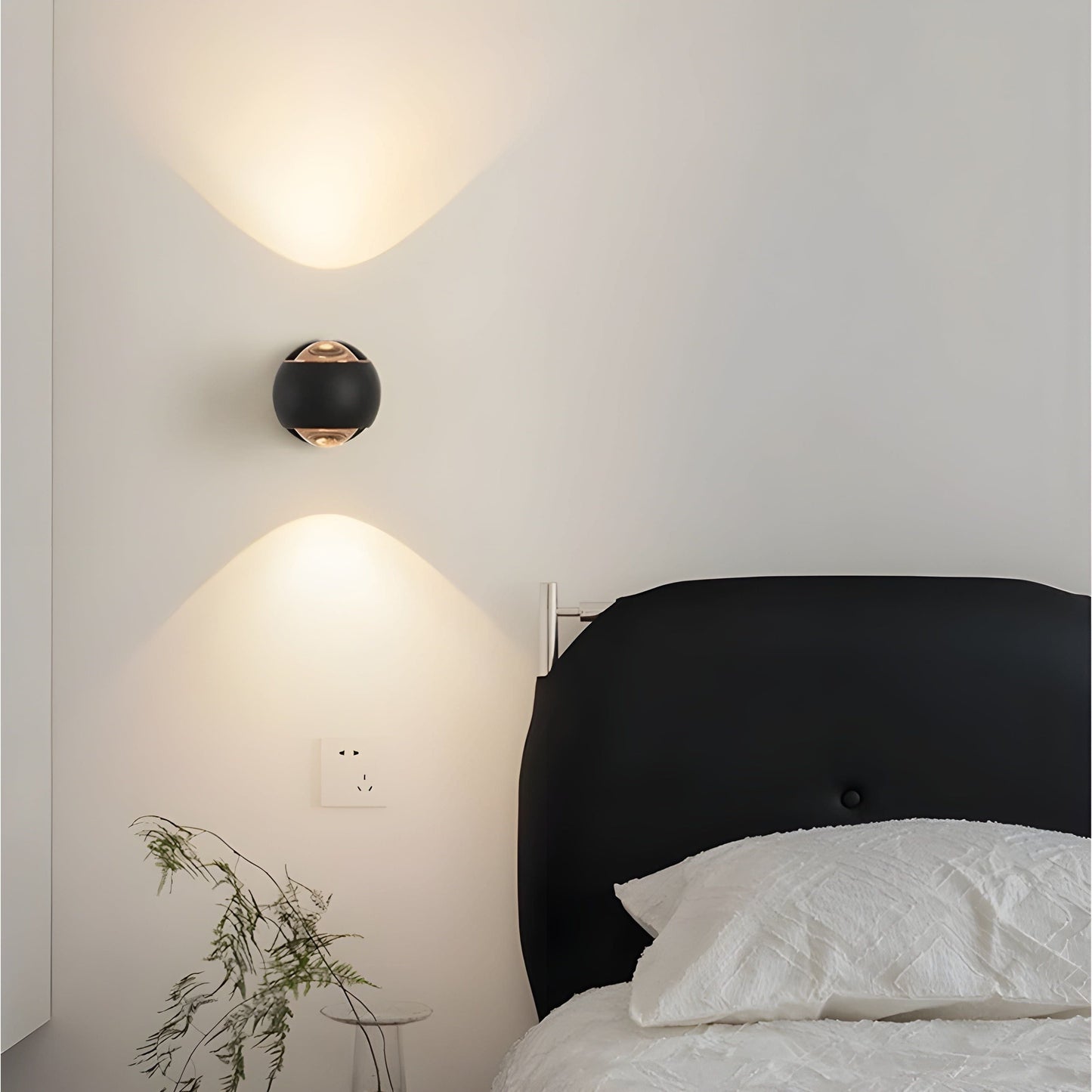 Black spherical Fitlybuy ModernEdge rotating LED wall light mounted above a bed, casting warm up-and-down light beams.