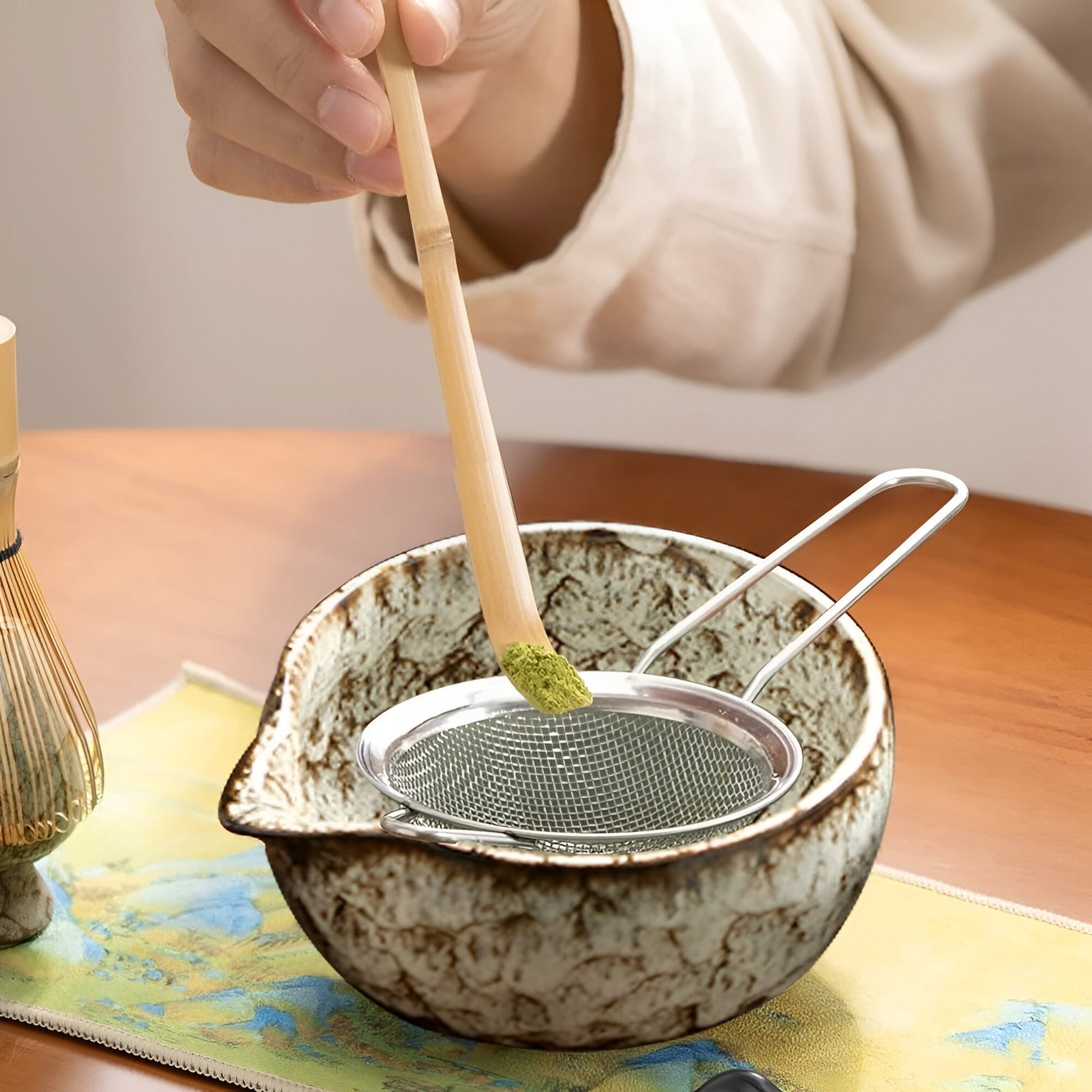 Fitlybuy matcha set: Hand holds bamboo scoop to sift green matcha powder into a rustic, textured ceramic bowl.