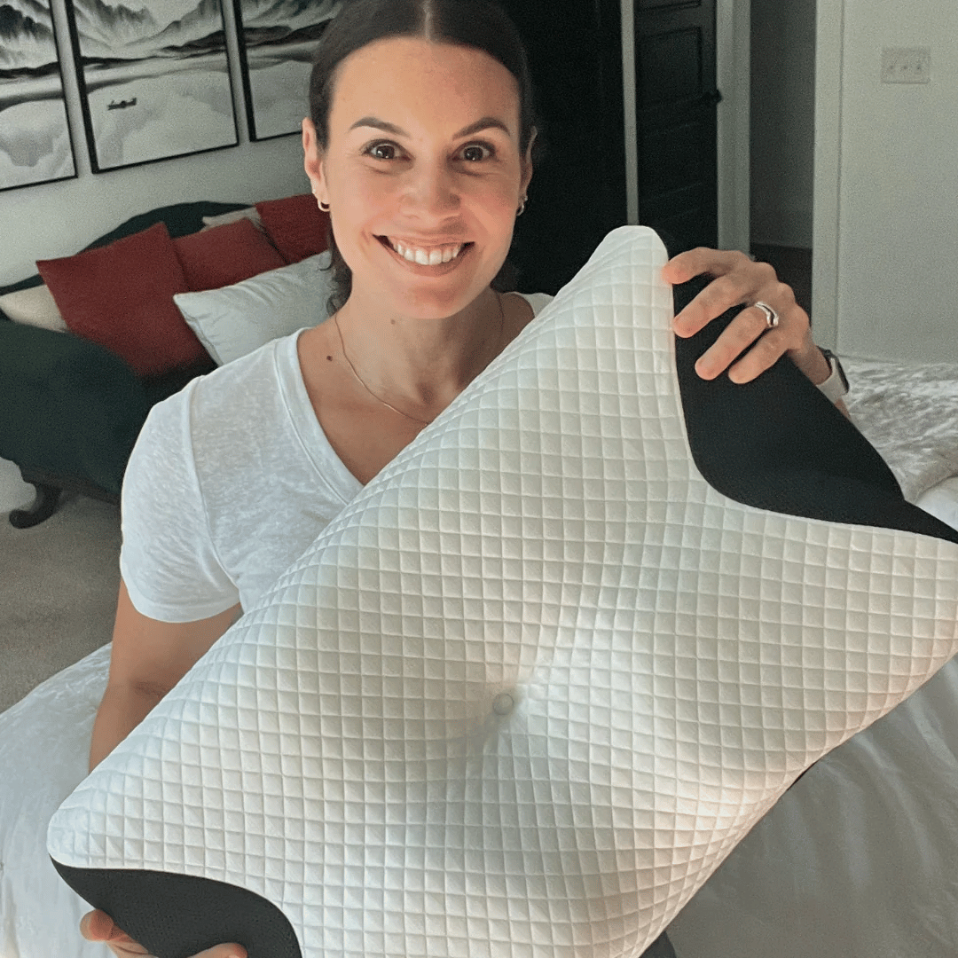 A woman holds a Fitlybuy white, quilted neck therapy memory foam pillow with black side accents in a bedroom.
