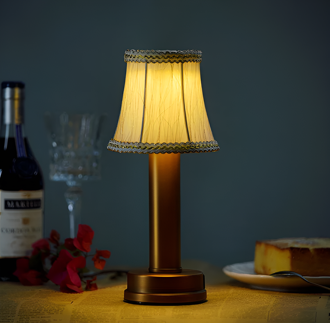 Vintage gold aluminium table lamp with a fabric shade, emitting warm LED light on a table with wine and cake.