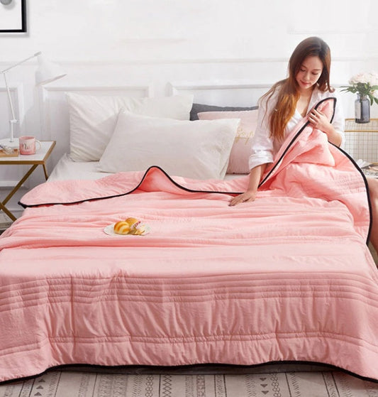 Woman sitting on a bed with a light pink, quilted Fitlybuy cooling blanket with dark trim.