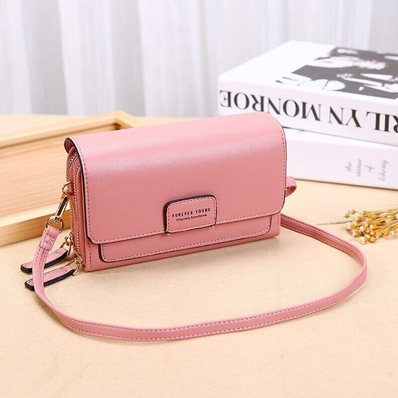 Dusty pink leather crossbody bag with a flap closure, gold zippers, and a “Forever Young” logo on a table.