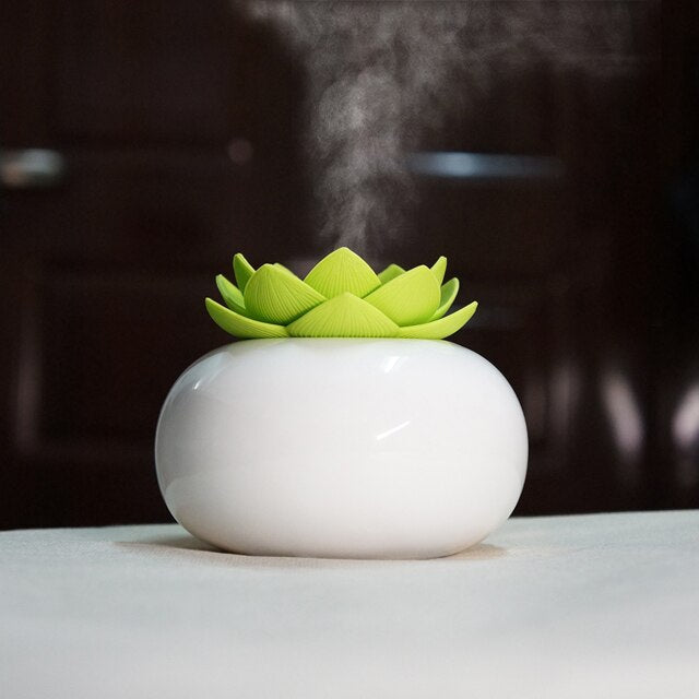 White ceramic lotus aroma humidifier with green petals releasing ultrasonic mist on a table.