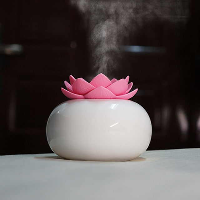 White ceramic lotus aroma humidifier with pink petals releasing ultrasonic essential oil mist on a table.