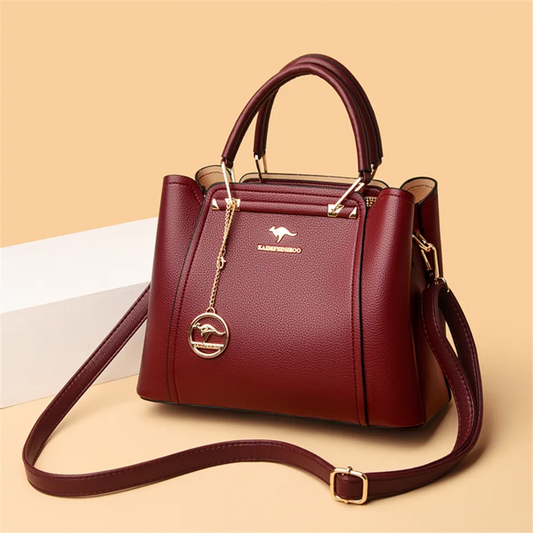 Burgundy pebbled PU leather handbag with gold kangaroo logo, charm, top handles, and adjustable crossbody strap.