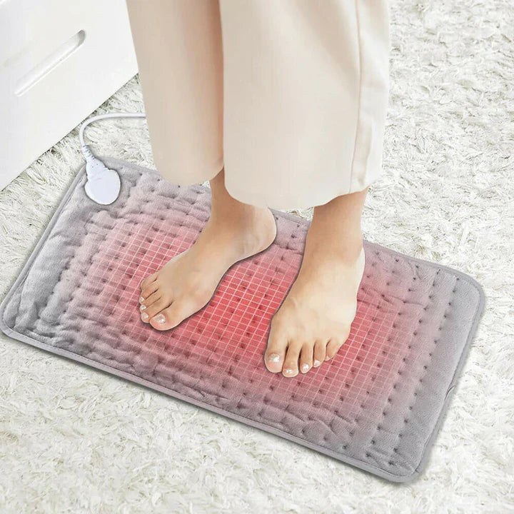 Person standing on a plush grey Fitlybuy electric heating pad with a glowing red grid design on a white carpet.
