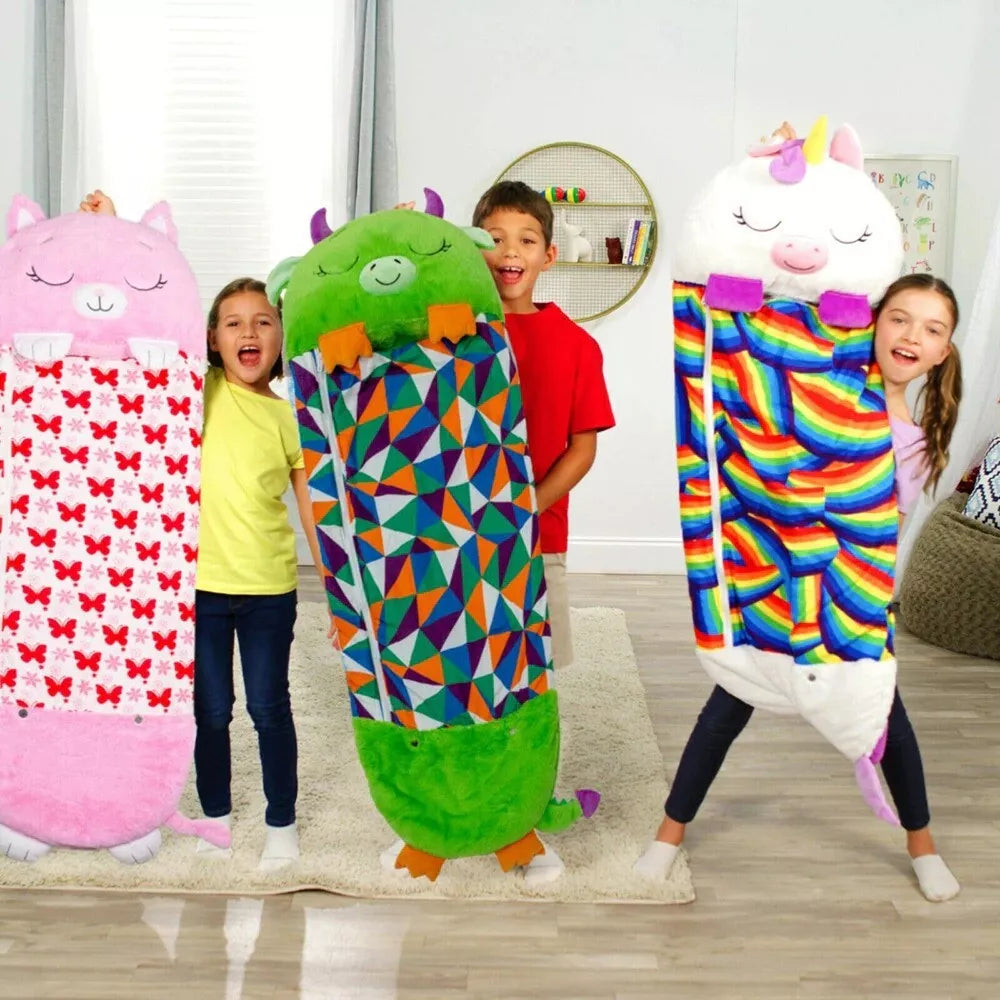 Three children stand in a room holding plush Fitlybuy SnugglePals sleeping bags: a pink cat, green monster, and unicorn.