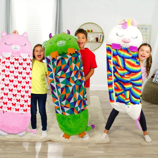 Three children stand in a room holding plush Fitlybuy SnugglePals sleeping bags: a pink cat, green monster, and unicorn.