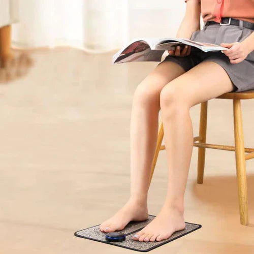 Person reading while feet rest on a black Fitlybuy EMS foot massage mat with blue electro-stimulation control device.