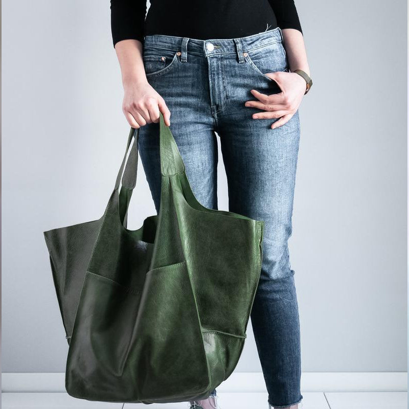 Person holding an oversized forest green leather weekender bag with structured handles, worn with jeans and black top.