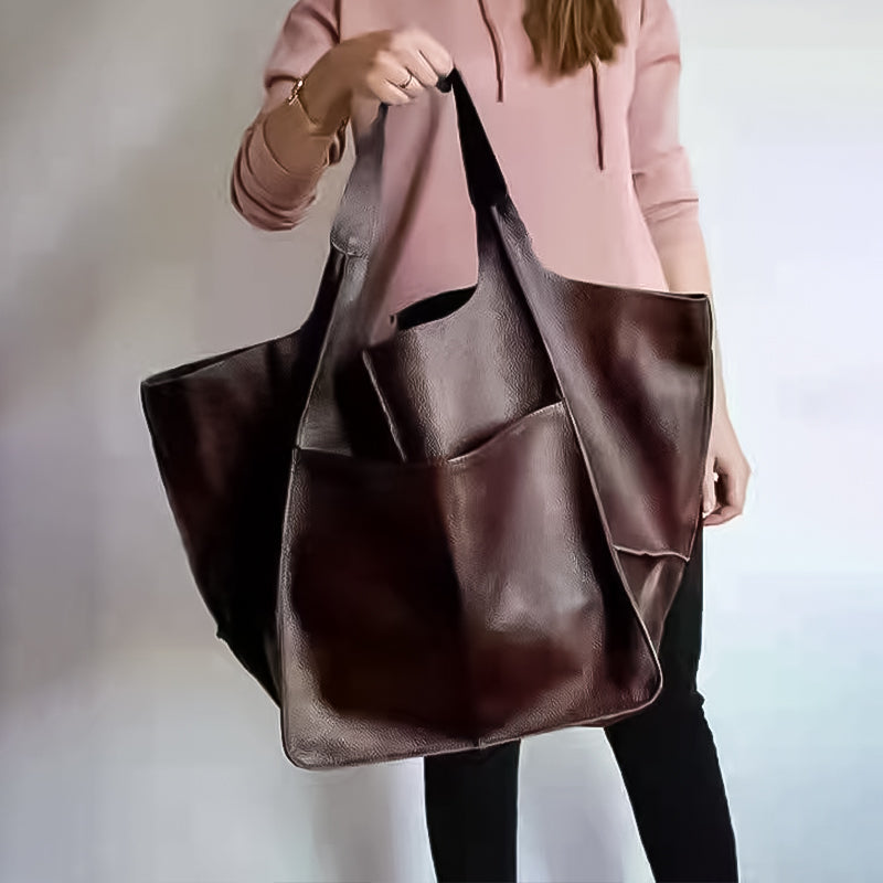 Person holds an oversized, dark brown leather weekender tote bag with a large external pocket and wide carry handles.